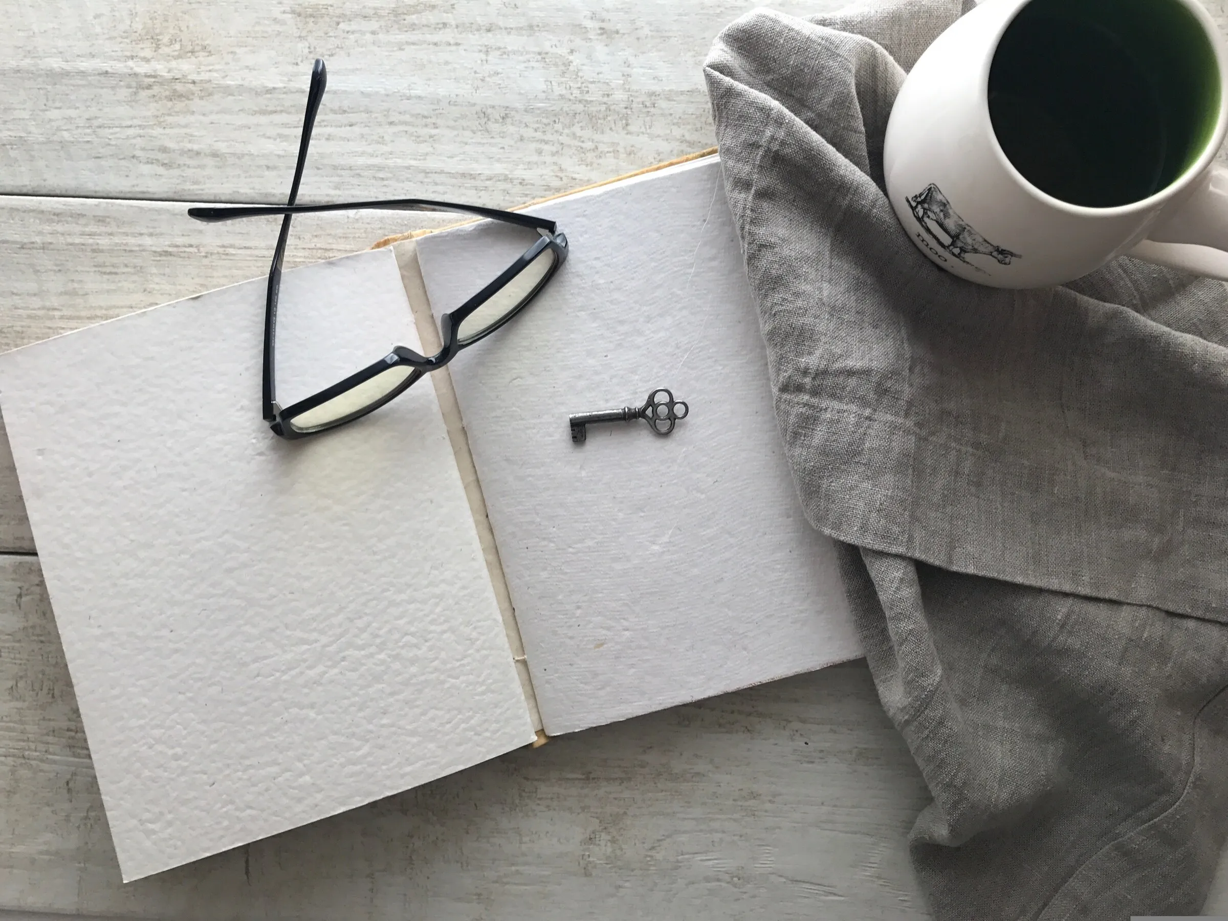 Open blank journal with reading glasses, a small key, and a cup of tea on a white wooden table