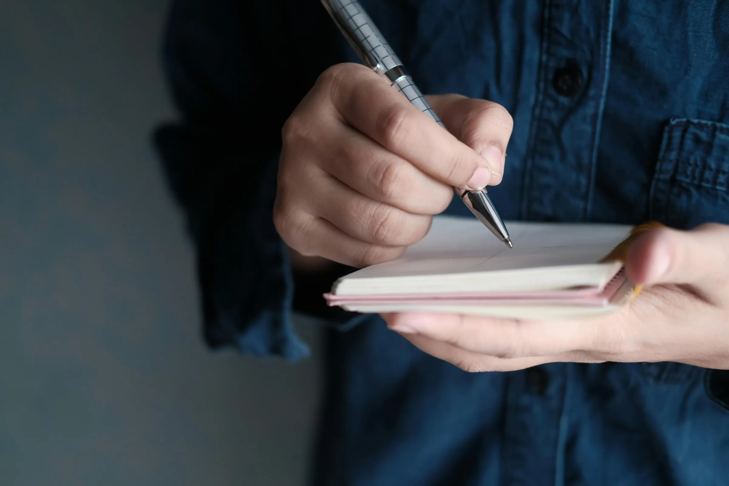 Close-up of a person in a denim shirt writing in a small open notebook with a silver ballpoint pen, capturing the quiet act of moving thoughts from mind to page