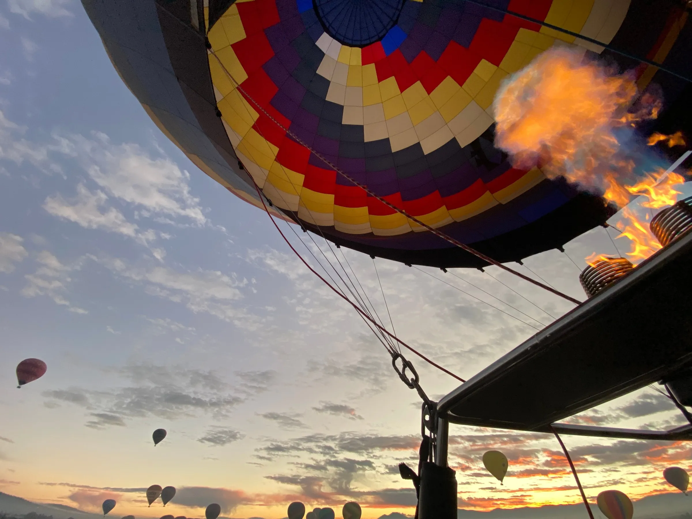 A vividly colored hot air balloon fires its burner against a soft sunrise sky, lifting off as other balloons drift over the valley below