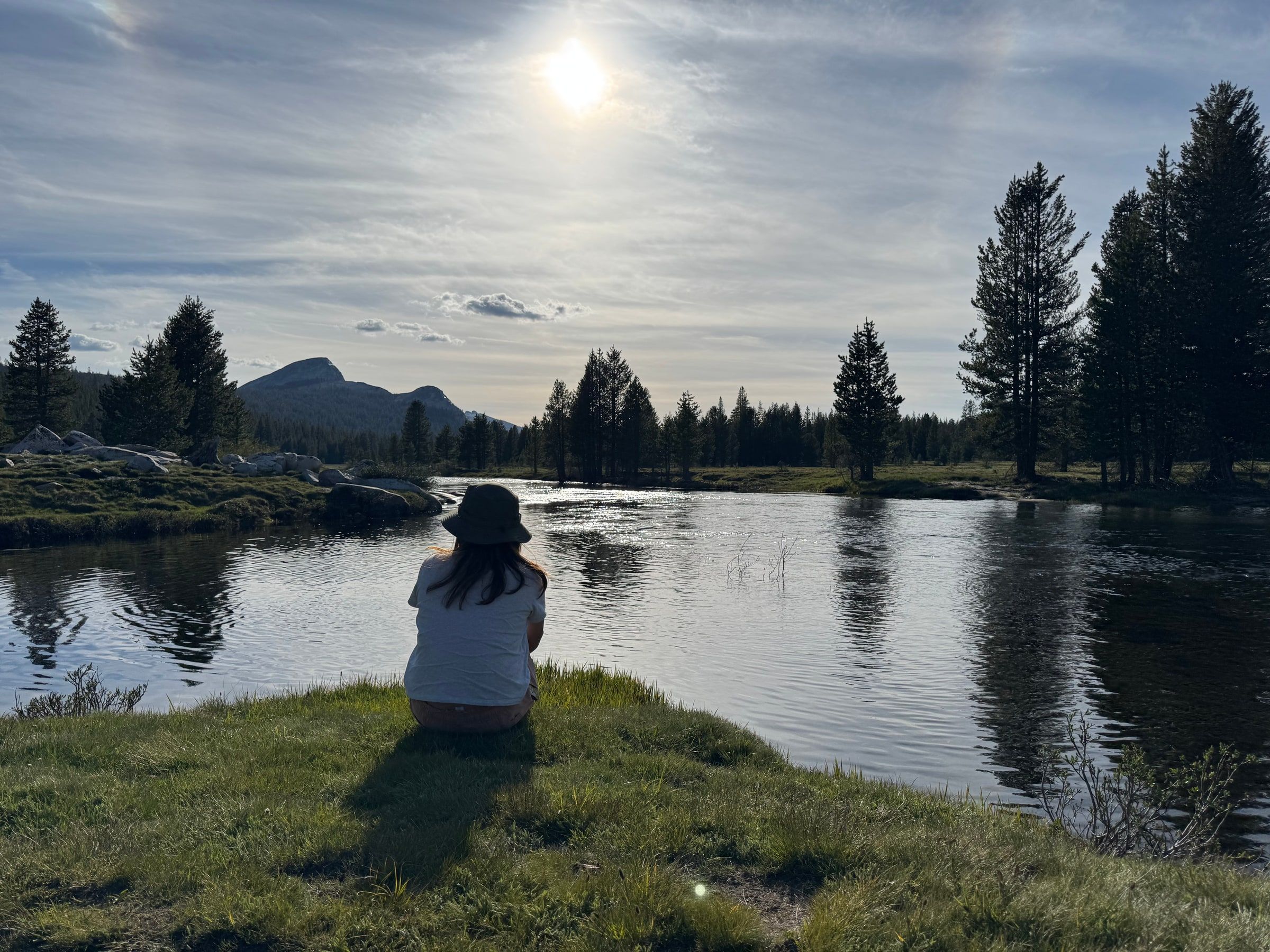 A person in a bucket hat sitting alone on a grassy riverbank at Tuolumne Meadows, Yosemite, quietly watching sunlight sparkle on the water with pine trees and mountains beyond